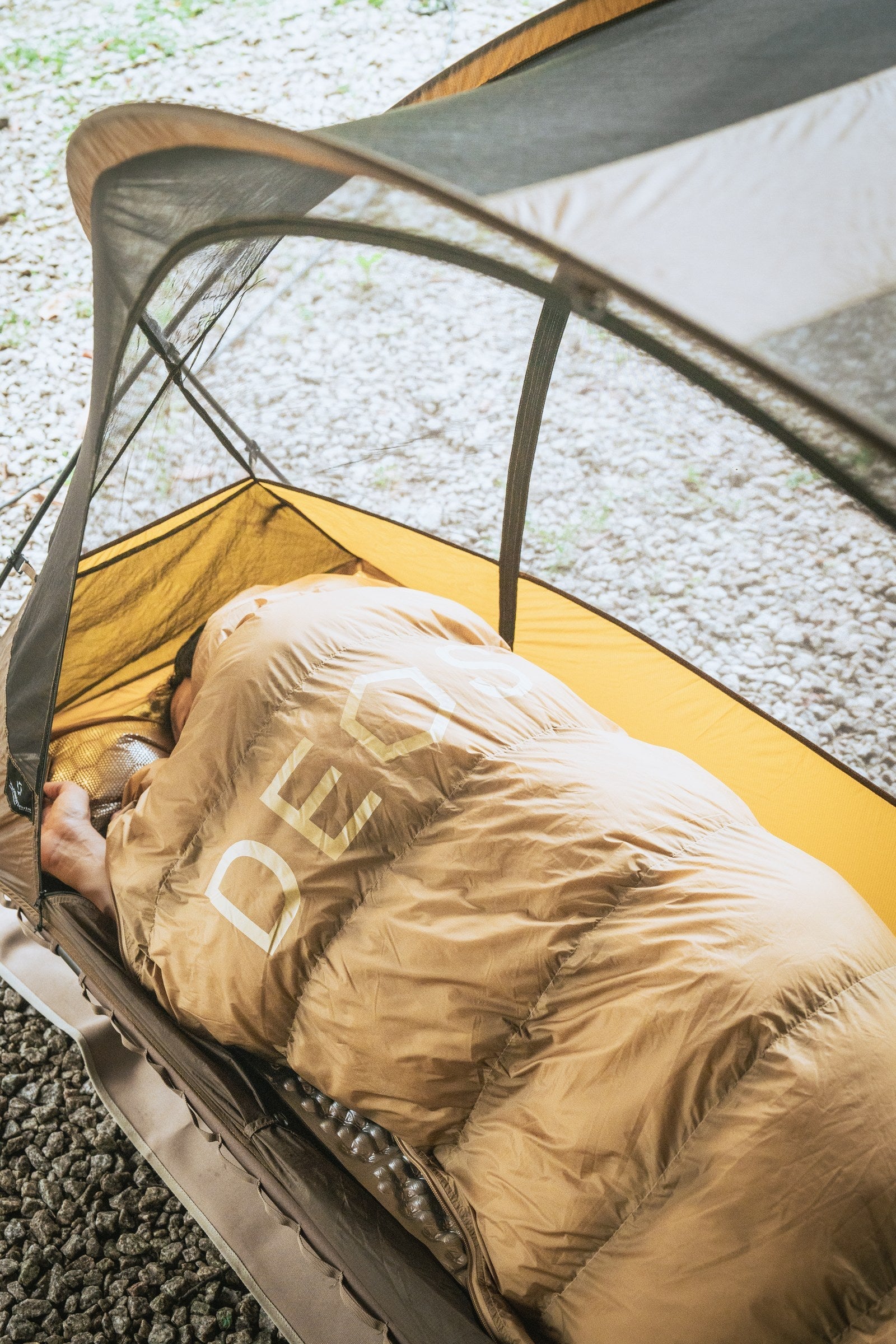 Camper resting on a cot under a tan DEOS camp comforter inside a tent