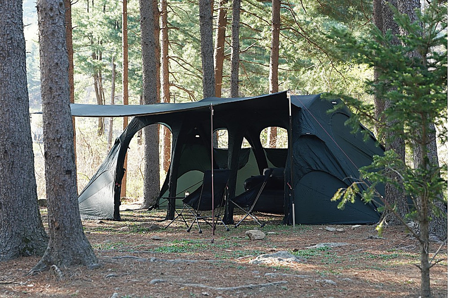 Black-coated T720+ side tarp extended as a front awning over camp chairs and a cot in a sunlit pine forest