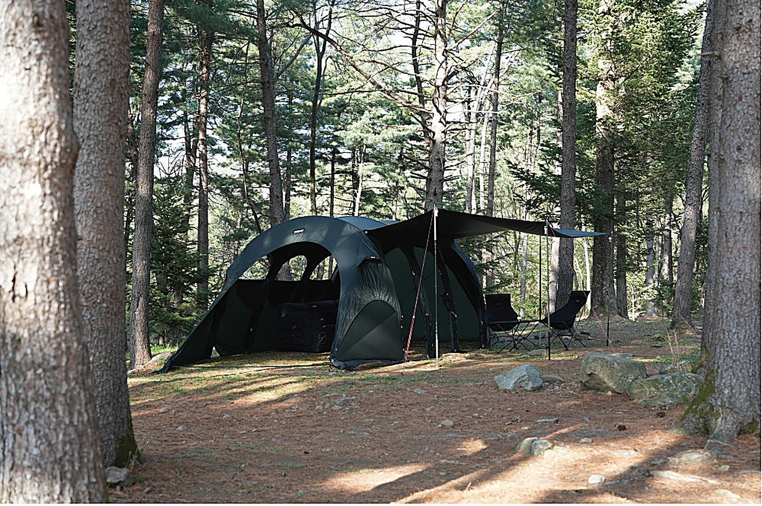 Gopamir T720+ tent with black-coated side tarp pitched among tall pines, camp chairs shaded beneath