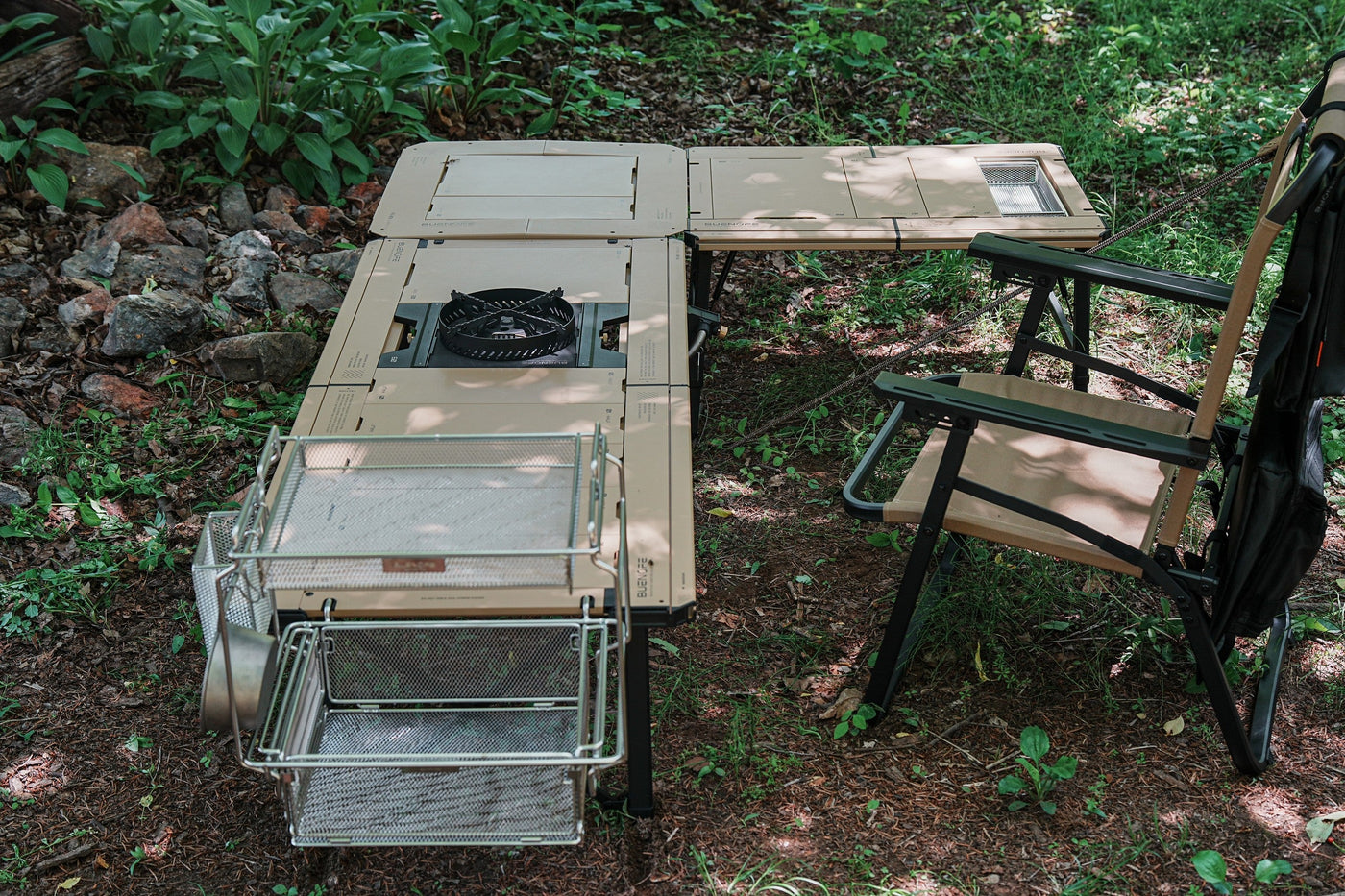 BUENOFE IGT Connect Table 1.5 extending a beige camp kitchen table into an L-shape outdoors, with a burner in the cutout and wire storage baskets and a hanger mounted on the accessory rail beside a camp chair.