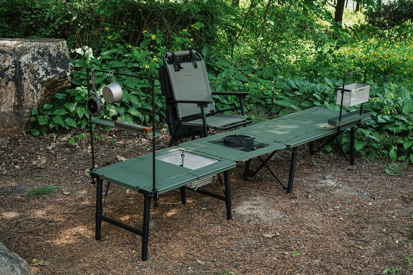 BUENOFE IGT Connect Table 1.5 extending an olive camp table into an L-shaped cooking station outdoors, with a hanging pot, wire basket and burner mounted along the accessory rail beside a camp chair.