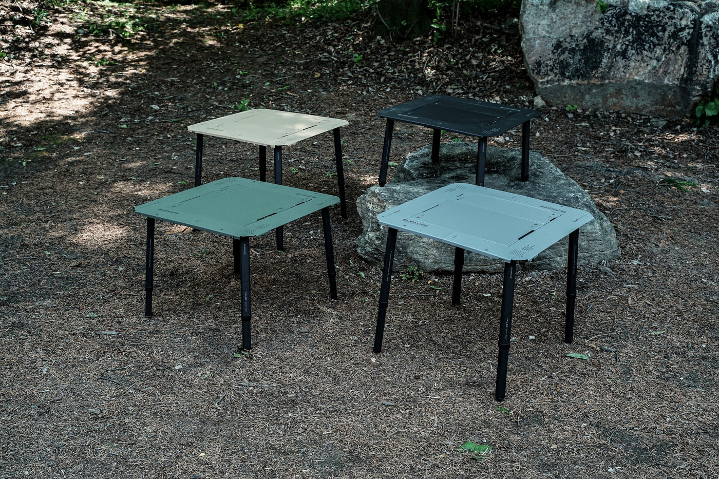 Four BUENOFE IGT Connect Table 1.5 standalone tables in tan, olive, silver-gray and black arranged together on a forest floor and rock, showing the full colorway range in an outdoor campsite.