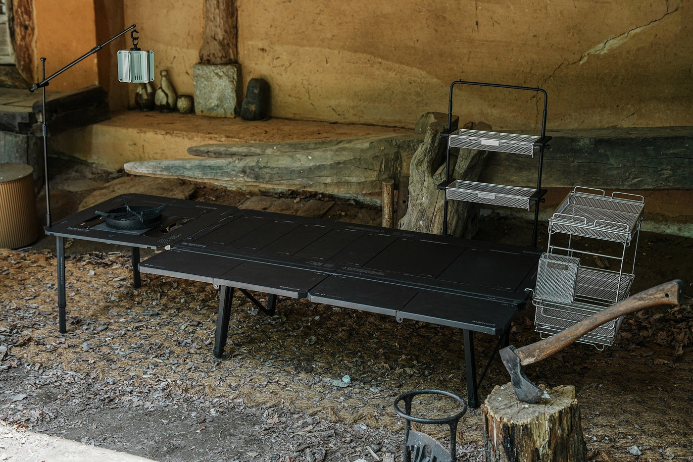 Editorial scene of a black BUENOFE IGT Connect Table 1.5 long table with a hanging lantern, wire racks and an axe on a stump, set against the rustic earthen wall of a traditional Korean house for a heritage-meets-outdoors mood.