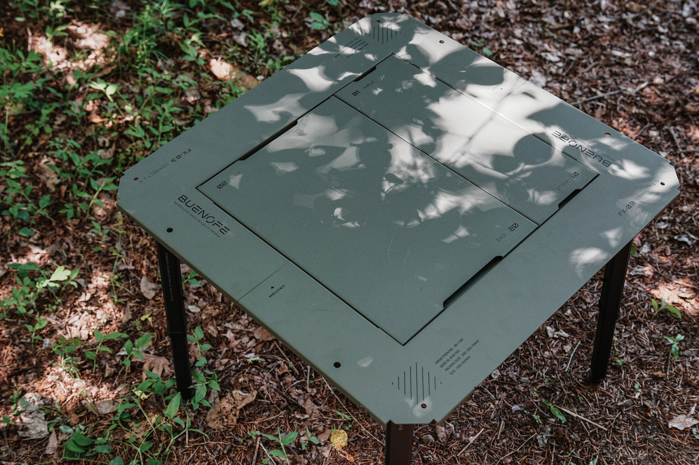 BUENOFE IGT Connect Table 1.5 assembled as an olive-green standalone table on a shaded forest floor, seen from above at an angle with soft dappled light.