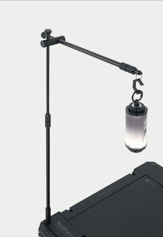 BUENOFE IGT Lantern Hanger photographed in a moody three-quarter studio scene, the pivoting boom arm angled down from the tall pole to hold a ribbed lantern over the edge of a dark tabletop, emphasizing the adjustable light direction.