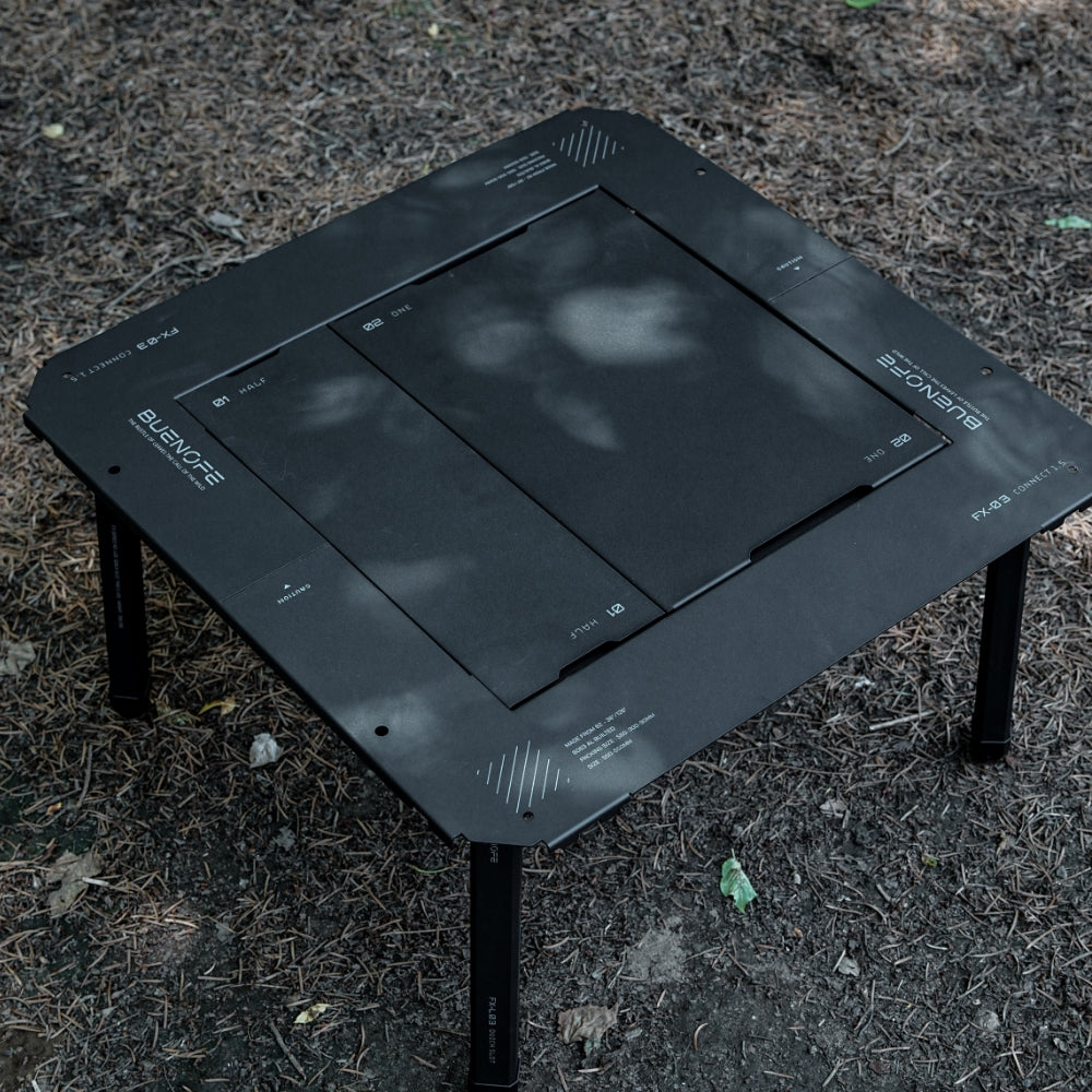 BUENOFE IGT Quick Slot Table Leg shown from directly above supporting an all-black IGT CONNECT 1.5 table on shaded forest ground, the black legs and dark tabletop panels forming a single-tone setup with English-only engravings.