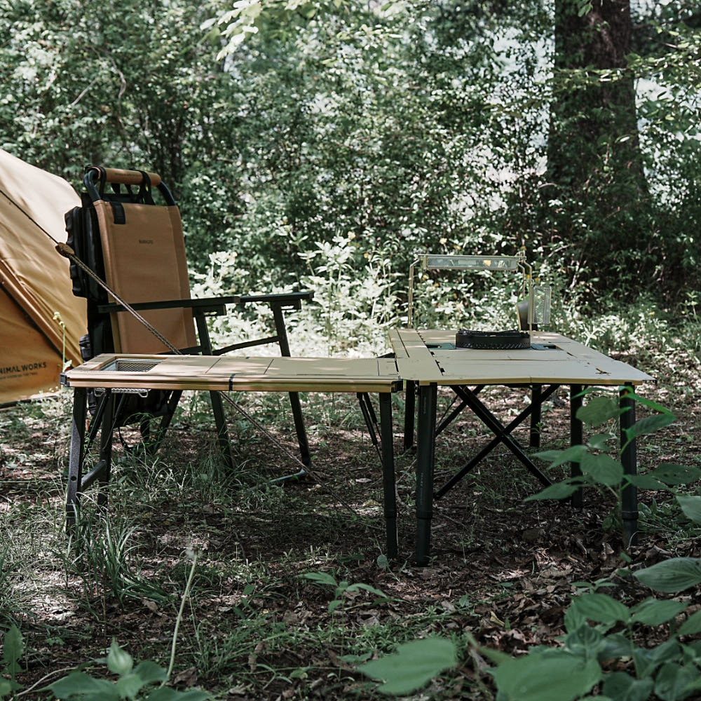 BUENOFE IGT Quick Slot Table Leg shown outdoors in a forest campsite, its black legs supporting two linked tan IGT tabletops extended in line beside a camp chair, tent and lantern for a larger shared cooking surface.