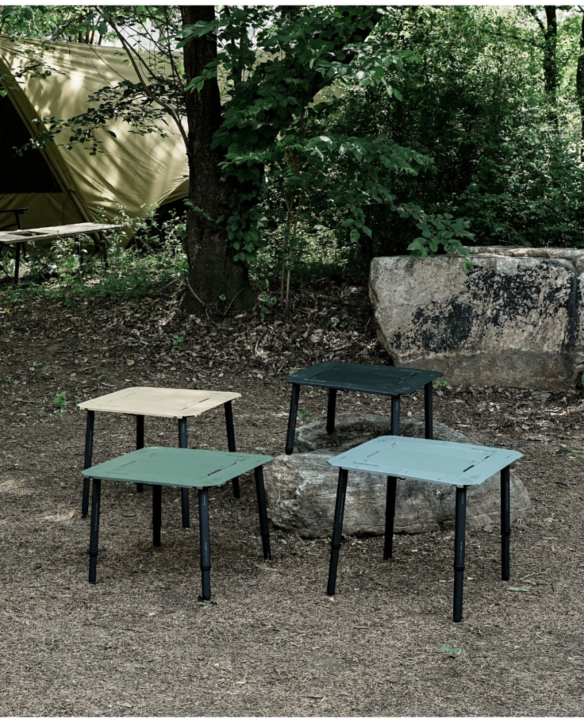 BUENOFE IGT Quick Slot Table Leg shown supporting four IGT tables in tan, olive, dark green and pale blue arranged on the ground of a wooded campsite near a tent and boulder, showing the legs paired with a range of tabletop colors.