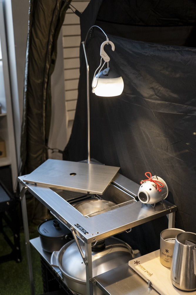 Close-up of the hook at the end of the arm, carrying a lit lantern above a camp kitchen shelf.