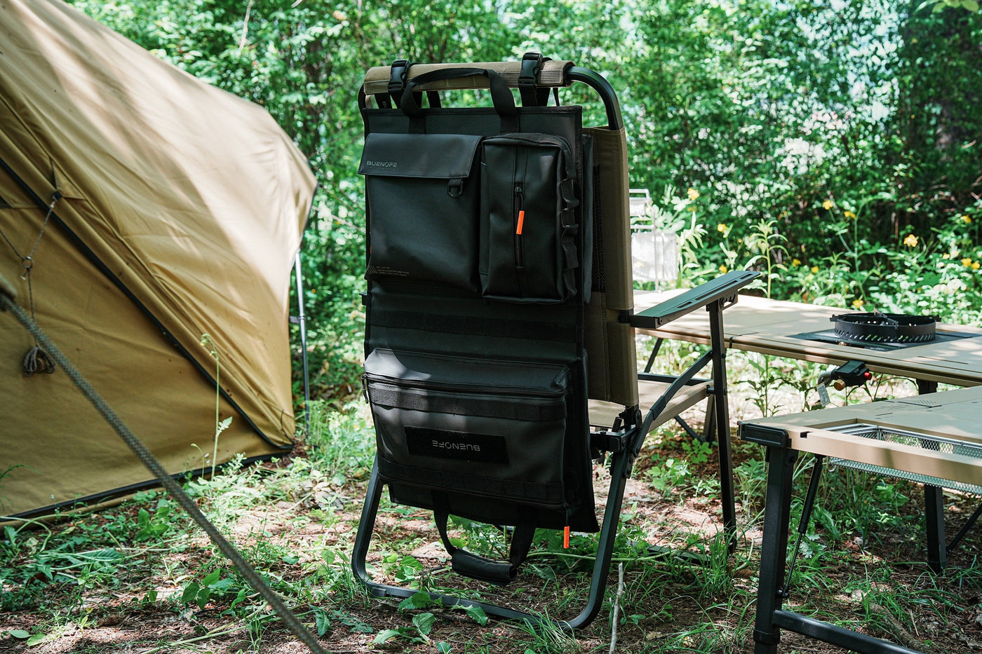 BUENOFE Multi Storage Bag Slim hanging on a camp chair back in a wooded campsite, wide landscape view with a tent, modular tables and a stove nearby, presenting the organizer as part of a full camp kitchen setup.