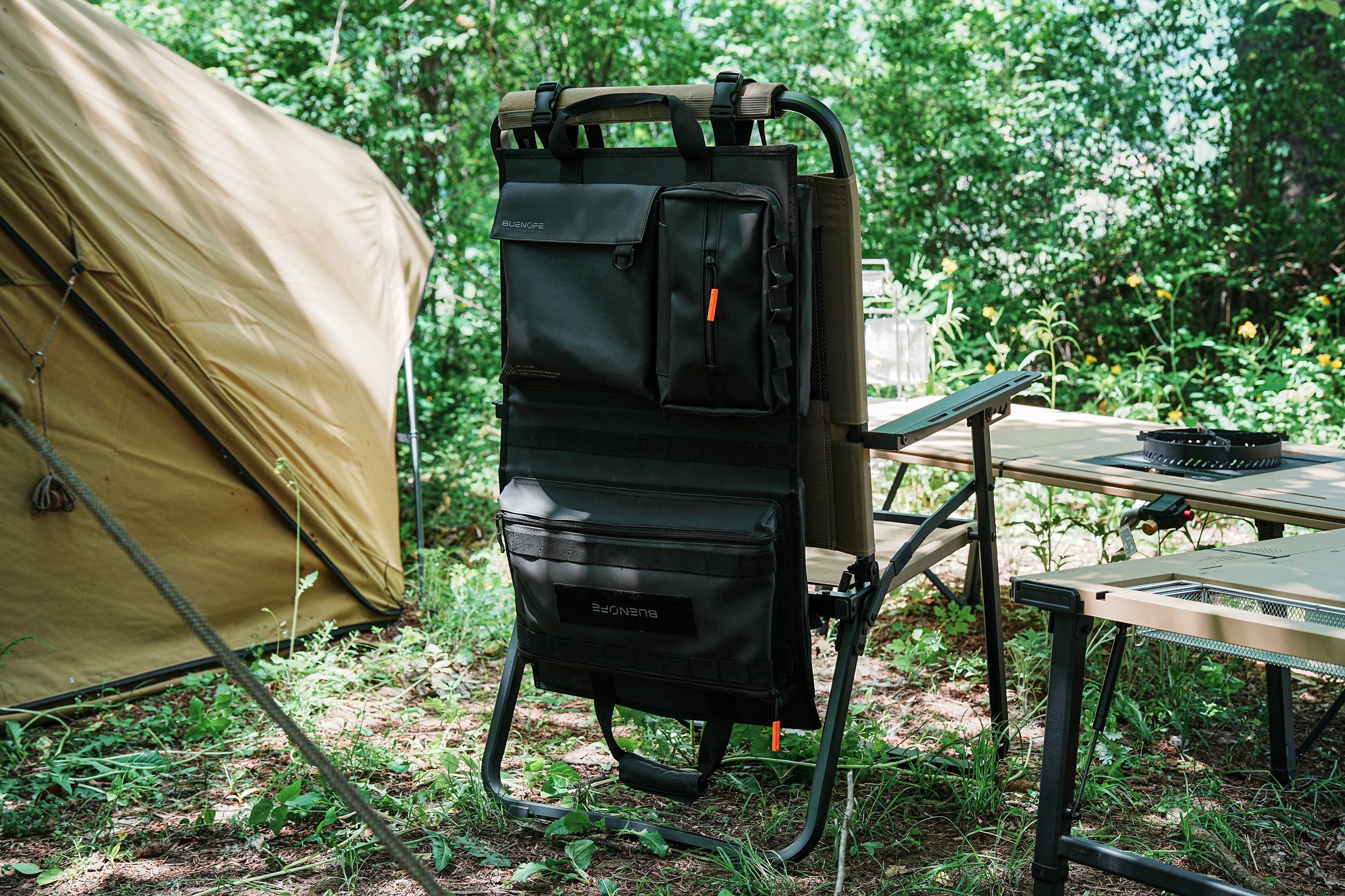 BUENOFE Multi Storage Bag Slim hanging on a camp chair back in a wooded campsite, wide landscape view with a tent, modular tables and a stove nearby, presenting the organizer as part of a full camp kitchen setup.