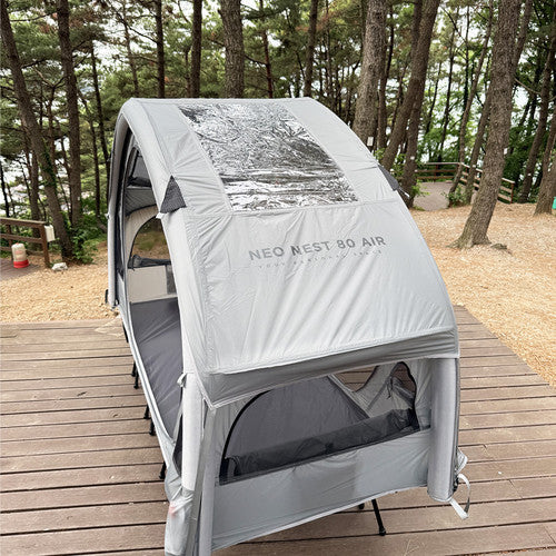 Neo Nest 80 Air with the included roof cover fitted, on a camp cot on a wooden deck under pine trees (cot sold separately).