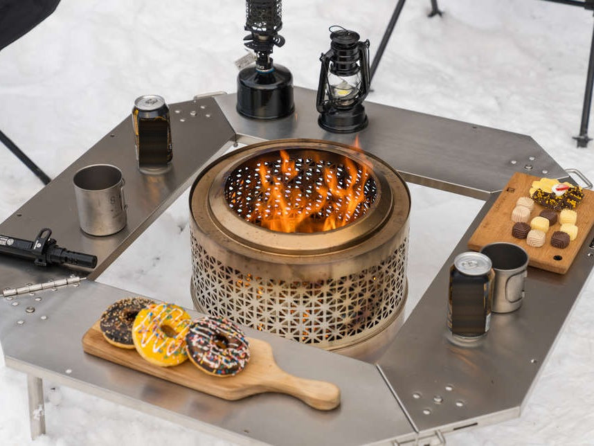 Wood-burning stove standing in the centre opening of the Biloc Octa table with flames going, seen from above with drinks and snacks on the surrounding plates. The stove is a separate purchase.