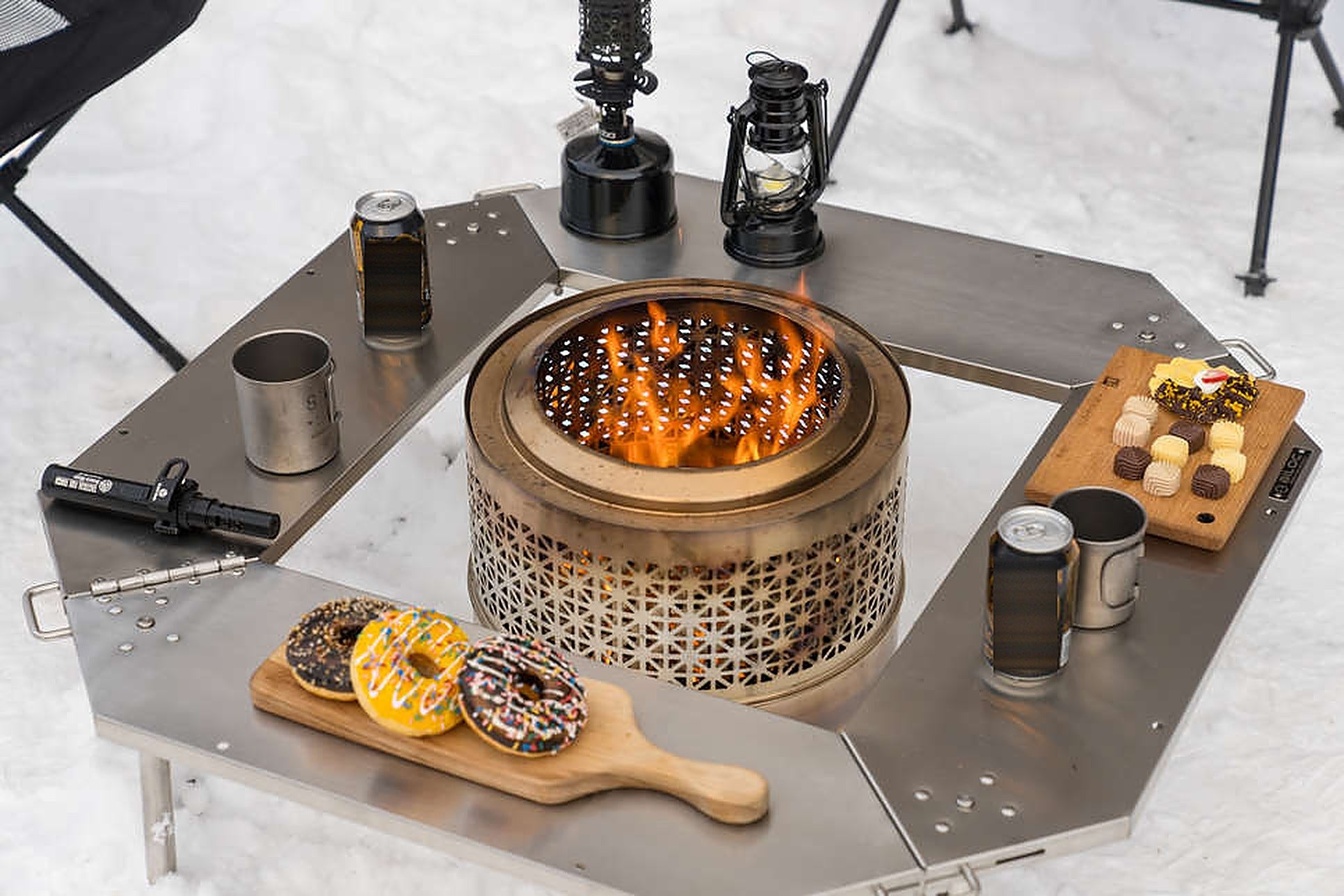 Top-down view of the Biloc Octa table ring with a wood-burning stove flaming in the centre opening, drinks and doughnuts on the plates. The stove is a separate purchase.