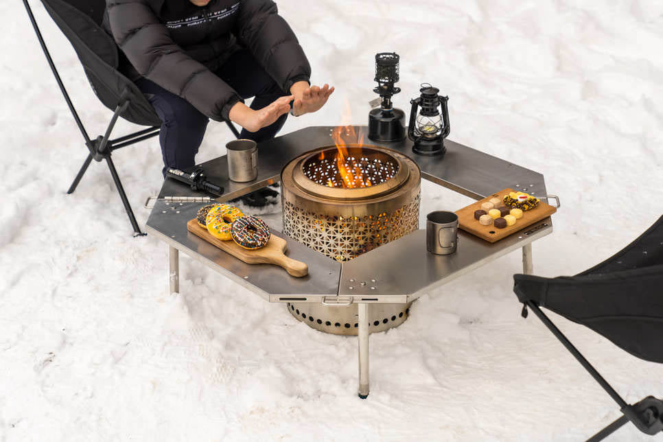 Person warming their hands over a fire burning in a stove set into the centre of the Biloc Octa table at a snowy campsite. The stove, lanterns and chairs shown are separate purchases.