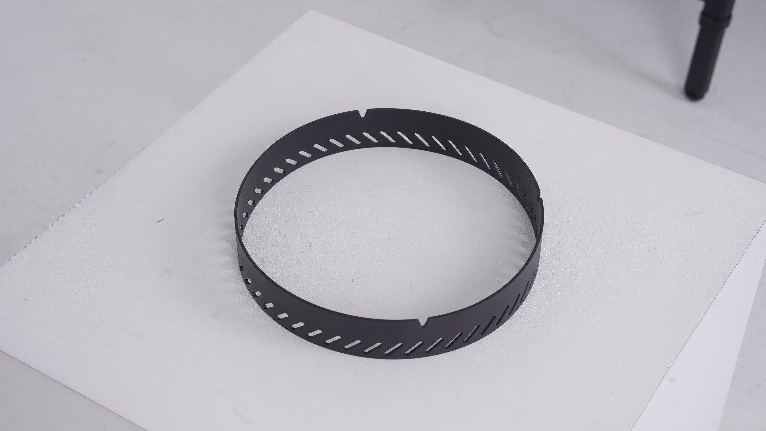 BUENOFE Stove Windscreen shown on its own as a matte black stainless steel ring resting on a white surface in a three-quarter view, highlighting the circular band and the row of angled diagonal air vents cut around its wall.