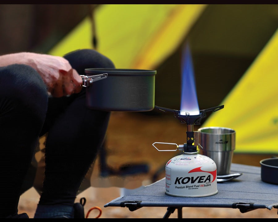 V1 Stove set up on a camp table with a pot and cup, lit and running