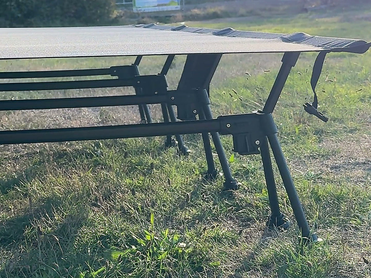 Wide Cot 80 in black set up on grass, low angle showing the taut sleeping deck and leg frames.